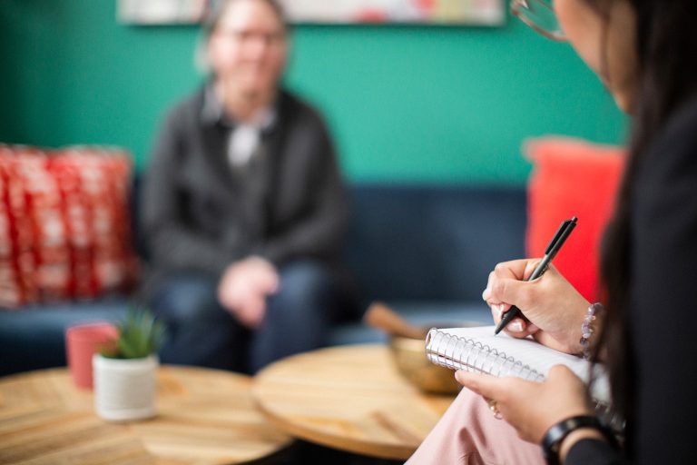In the foreground, a counsellor takes notes as a woman client sits on a couch in the background.
