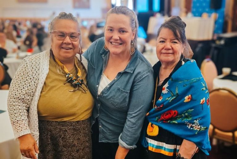 YWCA Edmonton CEO Rachel Foley (centre) poses with two Indigenous Elders in ribbon skirts in a conference room.