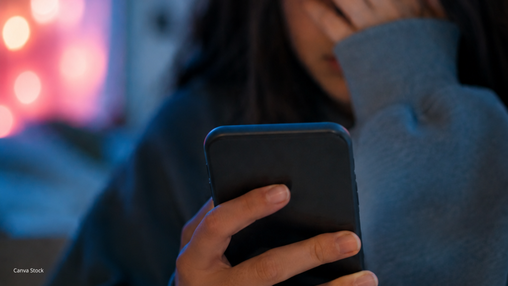A teen girl with dark hair covers her face as she looks at her smartphone.