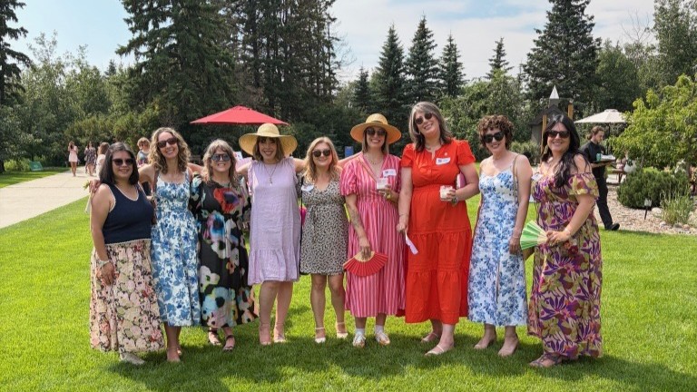 A group of women dressed in brightly coloured florals pose on the lawn of a greenspace with trees in the background