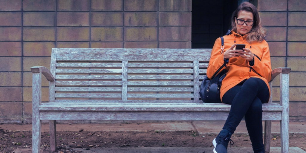 A middle aged woman wearing an orange jacket and dark rimmed glasses sits on a street bench reading on her smartphone.