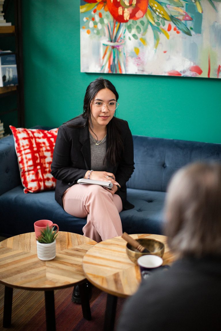 A dark-haired female counsellor wearing glasses sits on a couch in a colourful counselling space listening to a client whose back is to the camera in the foreground.