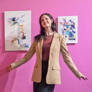 Artist Amy Polinsky poses in front of her artworks hung on a pink wall.
